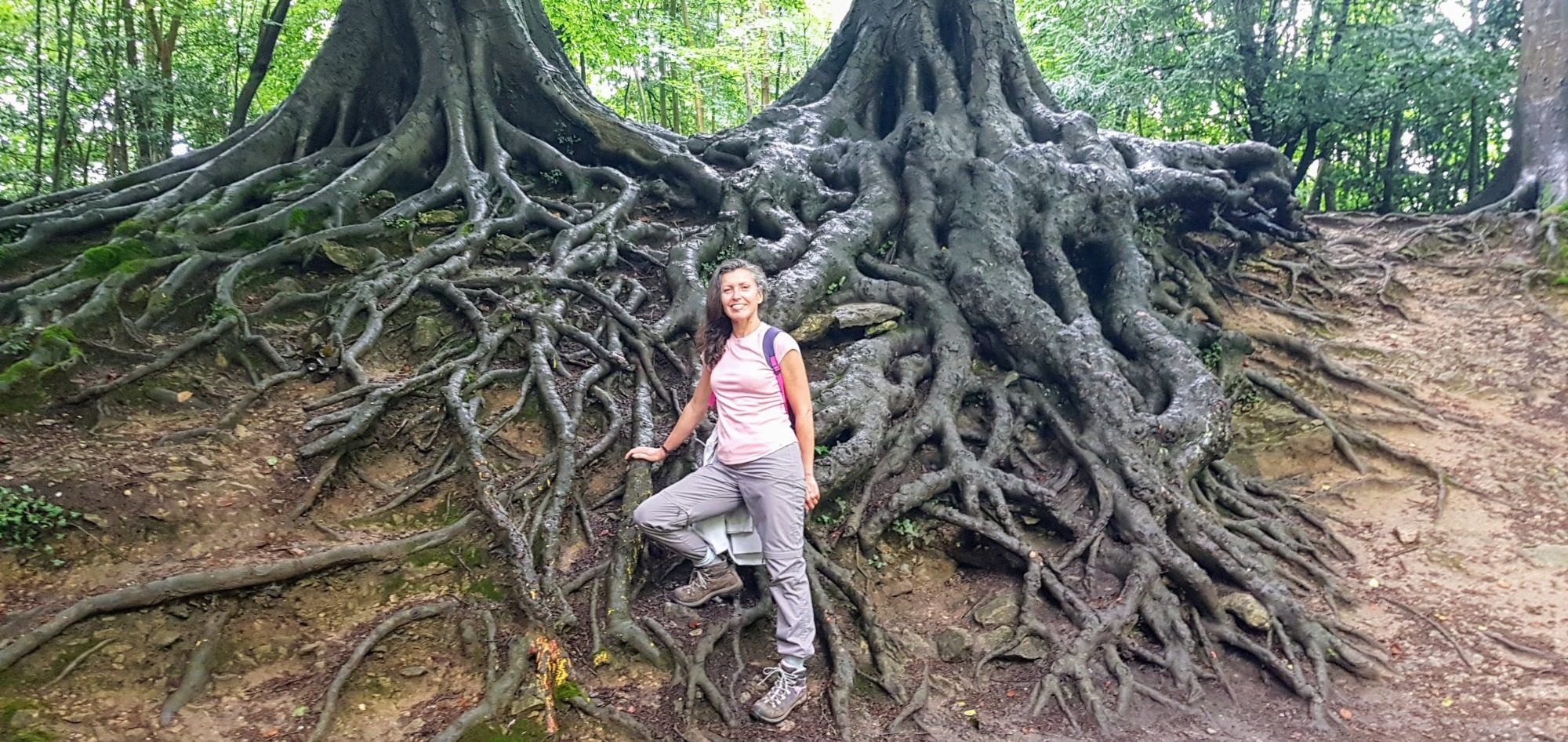 Miriam standing next to large, intricate tree roots in a forest setting.